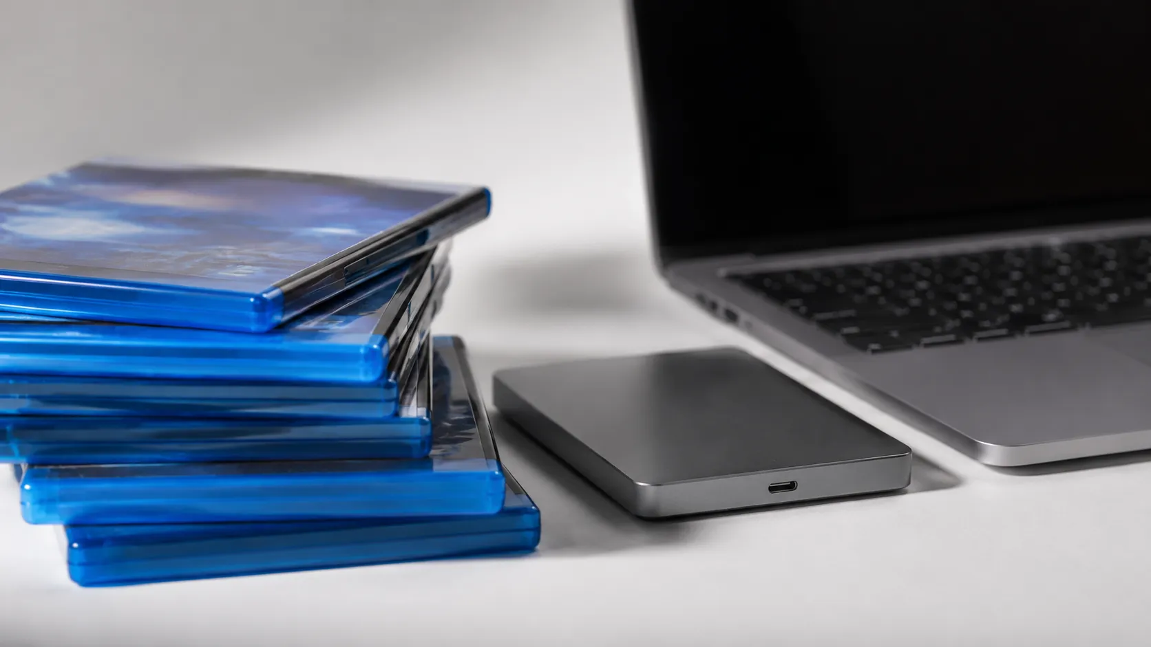 Stack of Blu-ray disc cases beside an external drive and MacBook ready for Blu-ray ripping on Mac